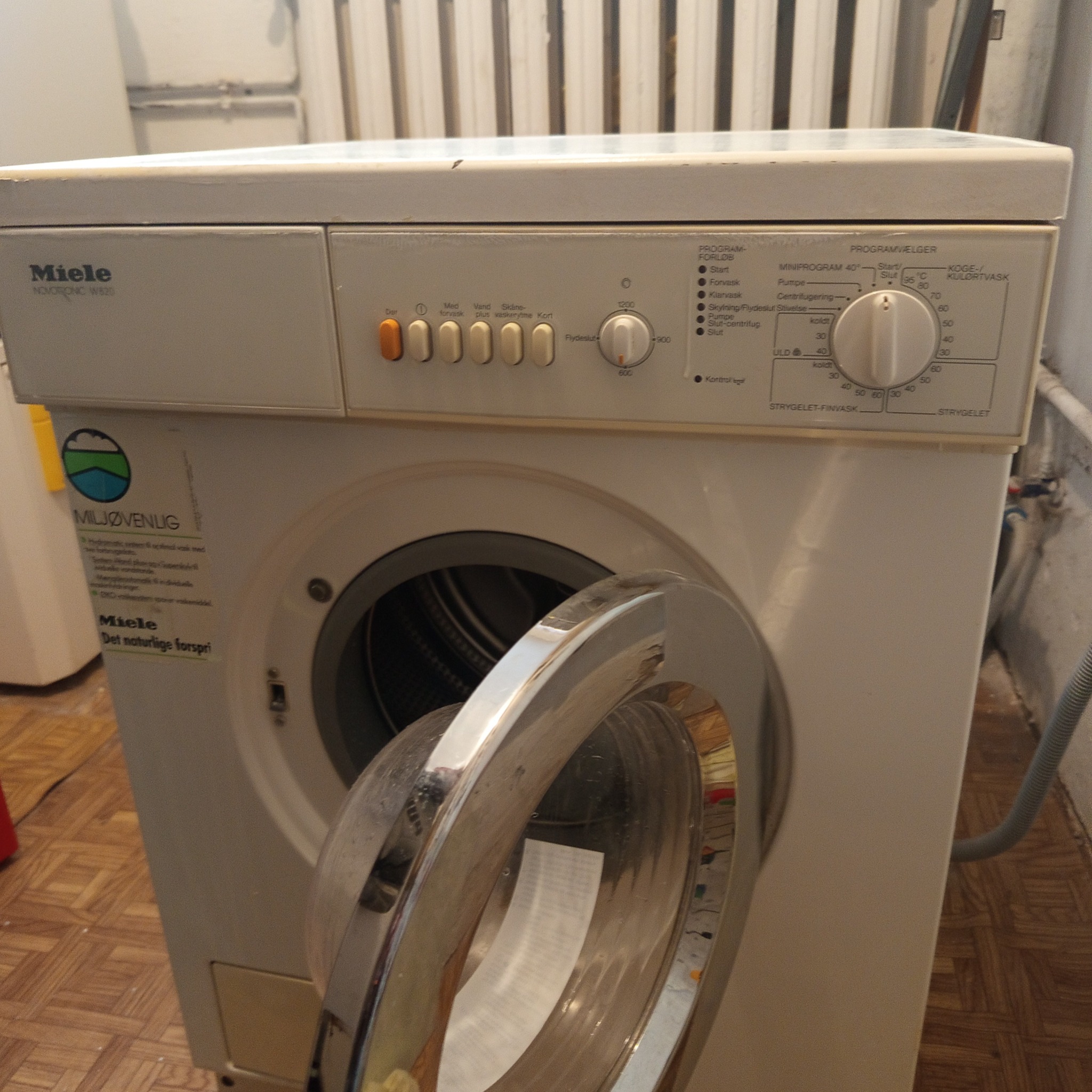 Otwarta pralka Miele z widocznym panelem sterowania i naklejkami informacyjnymi, stojąca na podłodze imitującej drewno. Widoczny wąż odpływowy.