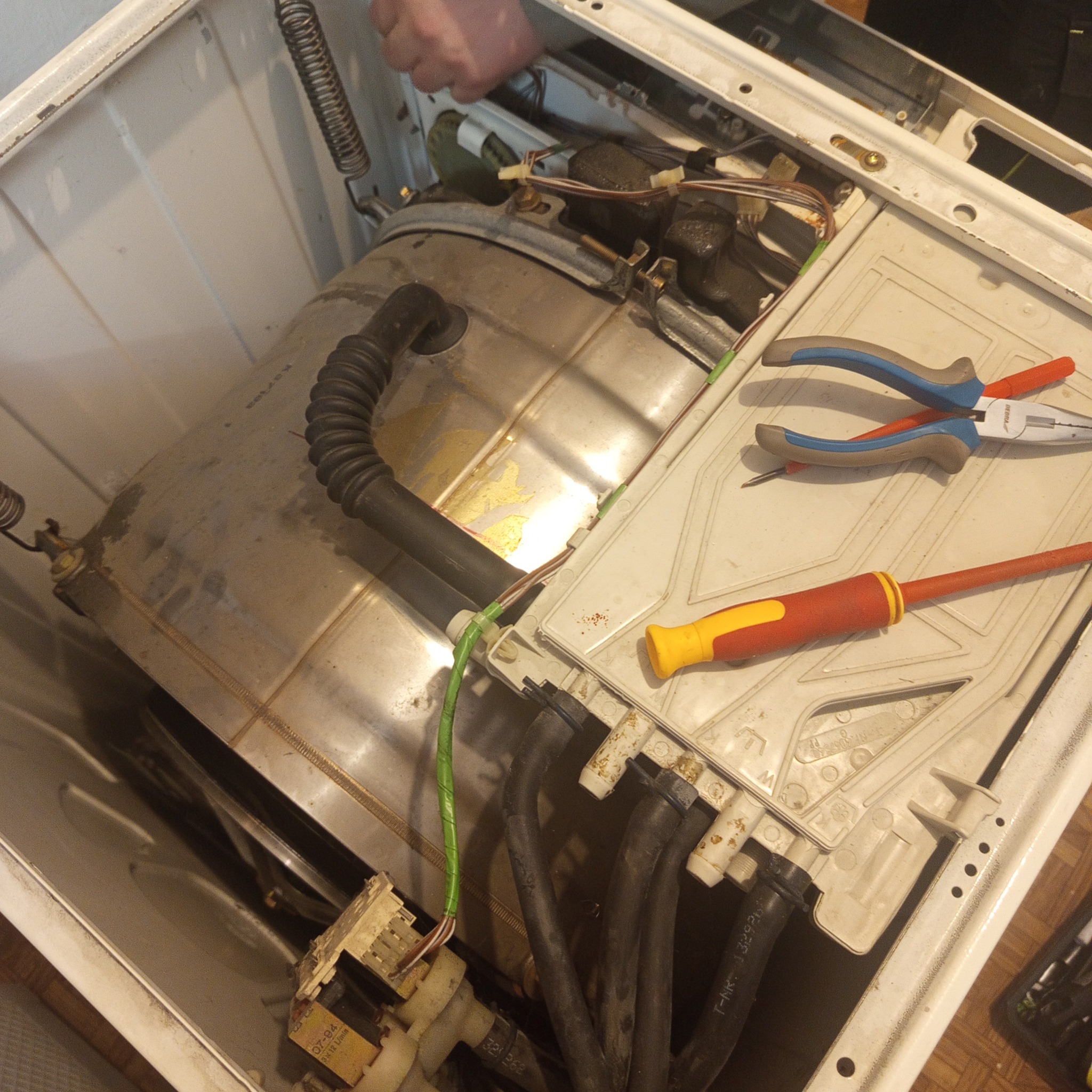 Wnętrze pralki w trakcie naprawy: bęben, przewody, narzędzia (kombinerki i śrubokręt). Widoczne elementy konstrukcyjne i podzespoły urządzenia. Ujęcie z góry, w trakcie serwisu.