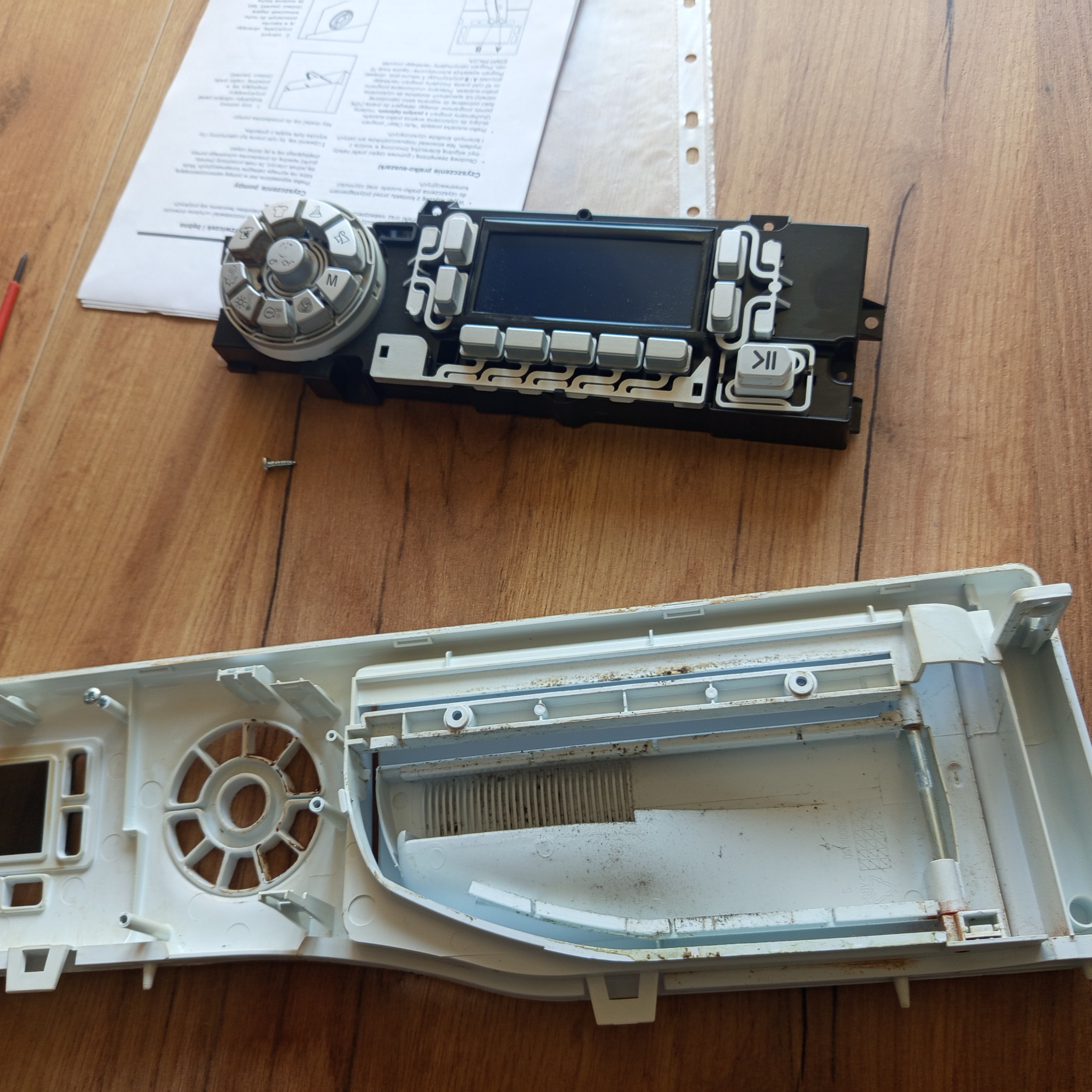 Rozłożony panel sterowania pralki z widocznym wyświetlaczem, przyciskami i pokrętłem, obok instrukcja i śrubokręt, na drewnianym blacie. Widoczne ślady użytkowania.