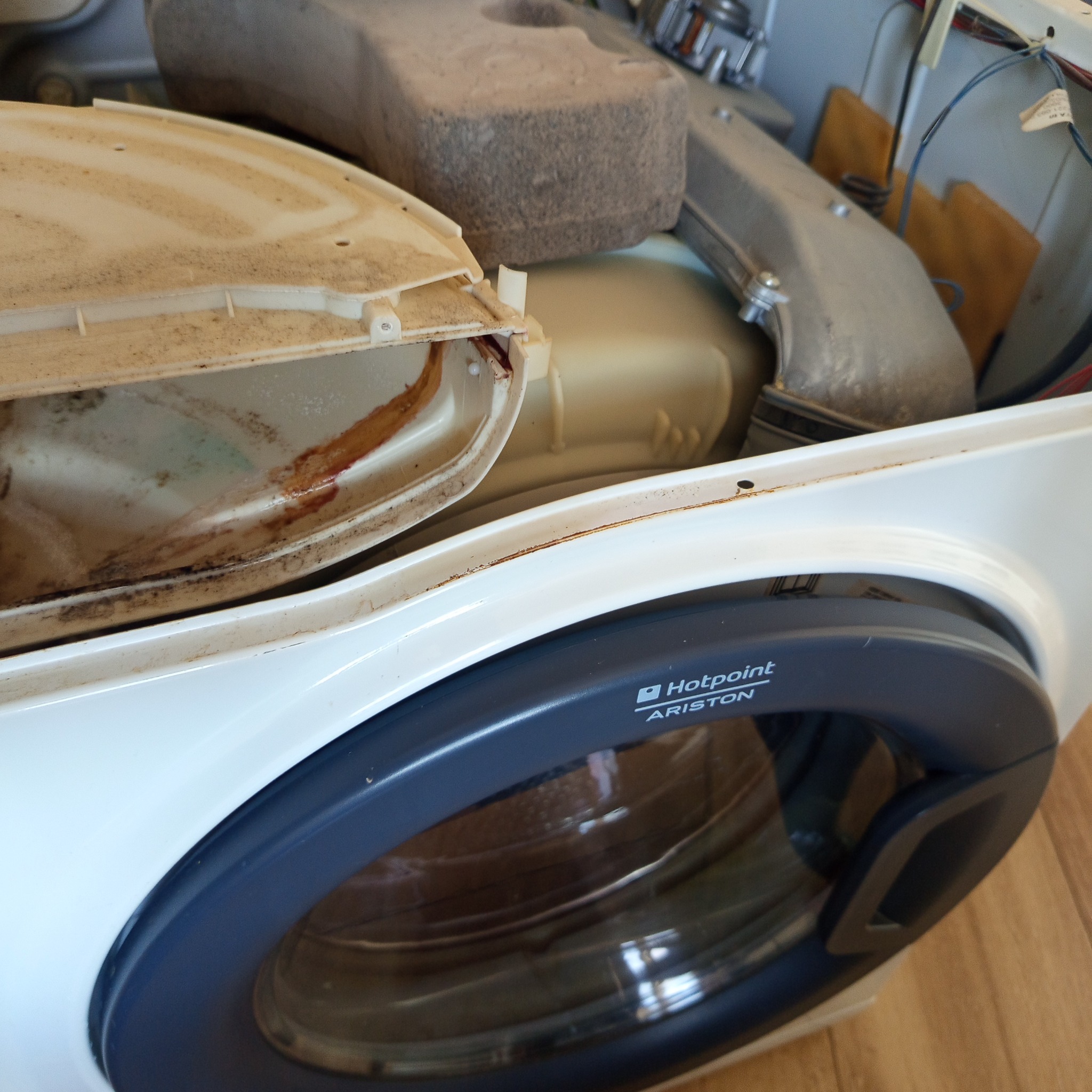 Wnętrze rozmontowanej pralki Hotpoint Ariston, widoczne zabrudzenia i osady. Ujęcie z bliska na elementy urządzenia w trakcie naprawy lub czyszczenia.