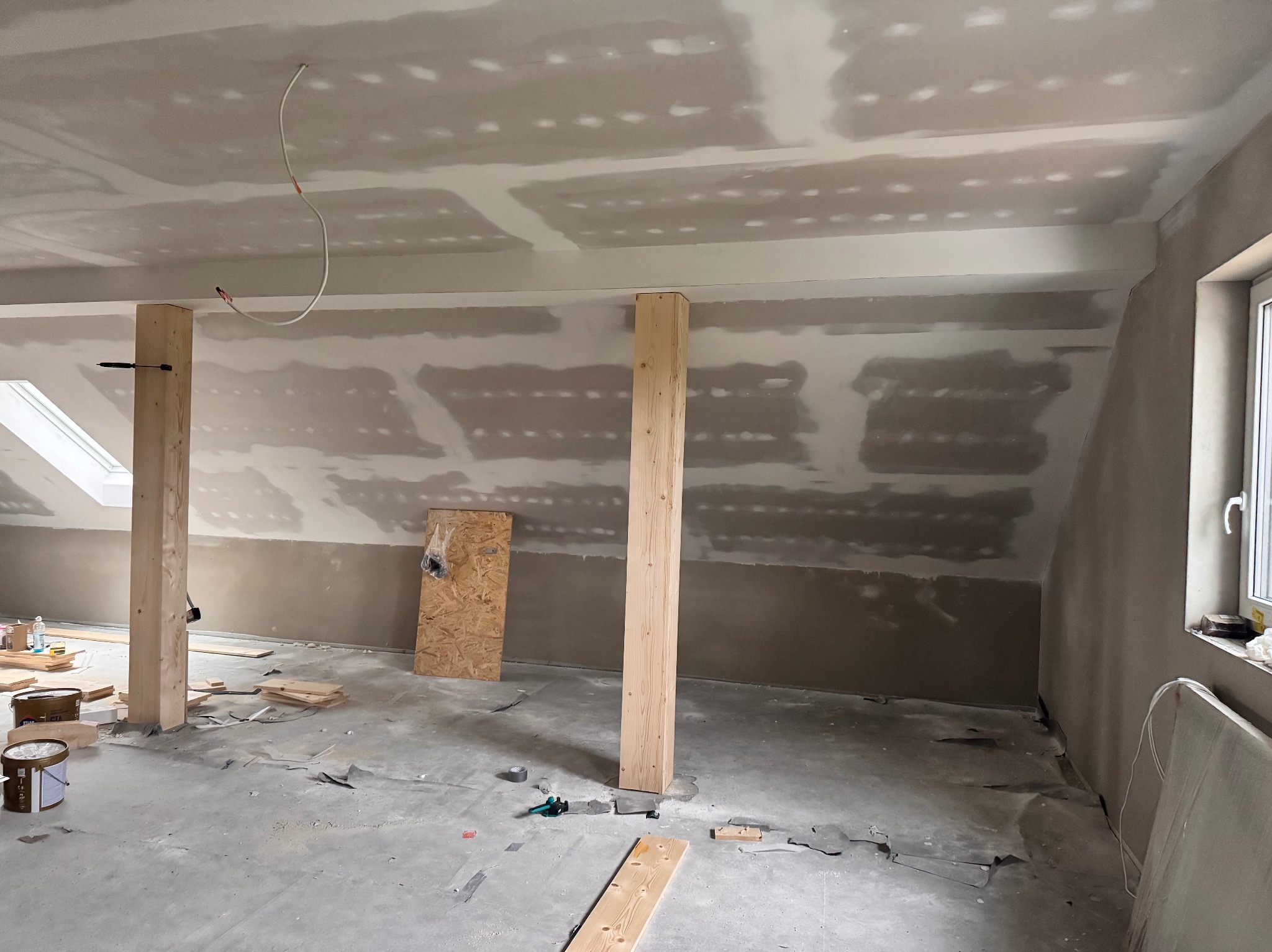 Poddasze w trakcie prac wykończeniowych: płyty gips-karton, drewniane podpory, widoczne narzędzia i materiały budowlane. Okno dachowe i standardowe okno doświetlają wnętrze.