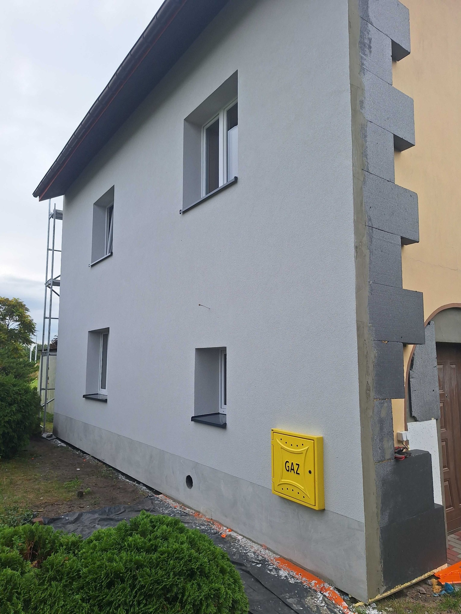 Jasnoszara elewacja domu z żółtą skrzynką gazową. Fragment ocieplenia z szarych płyt styropianowych na rogu budynku. Widoczne okna i fragment rusztowania.