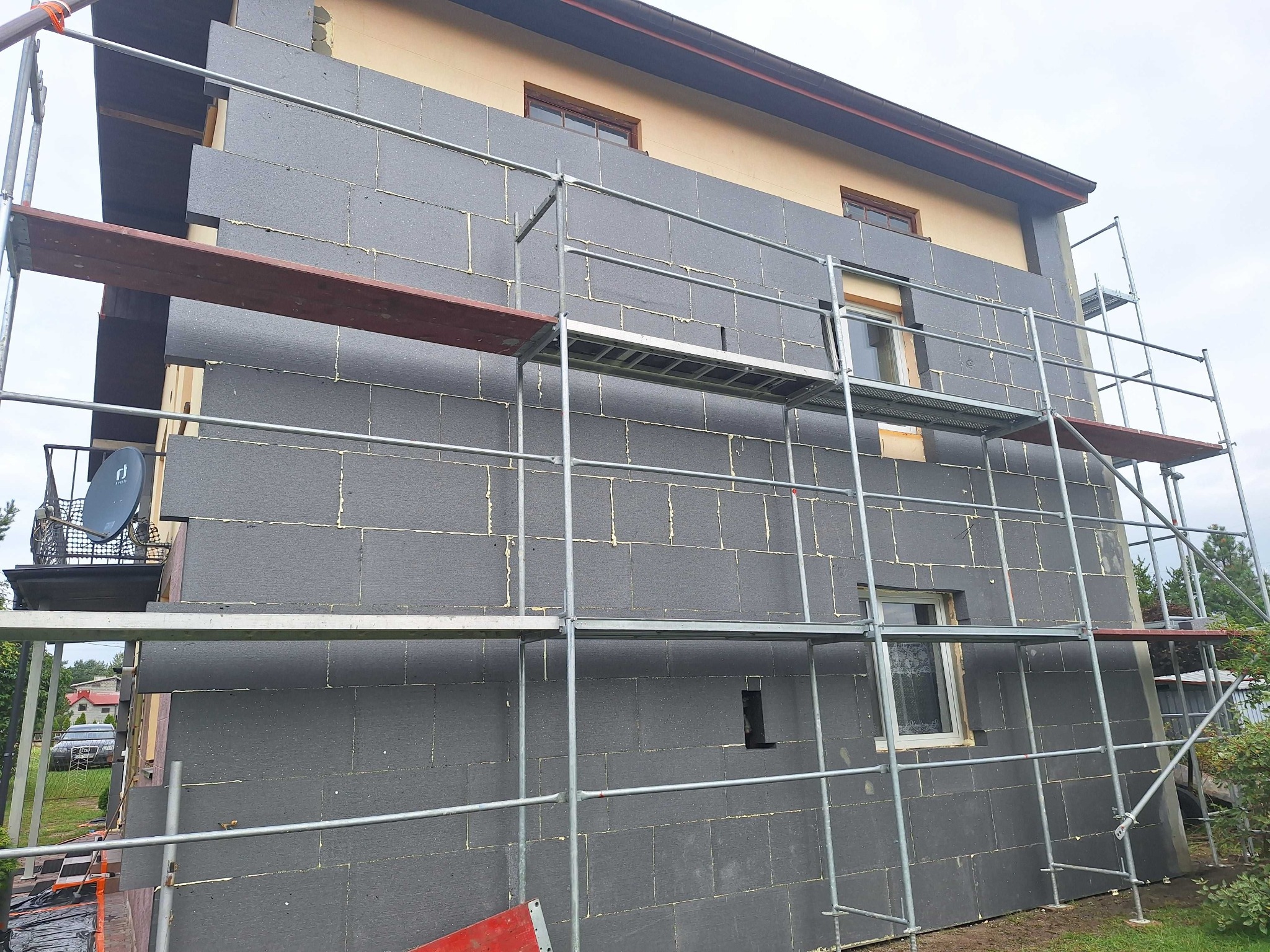 Dom w trakcie ocieplania: szare płyty styropianowe na elewacji, widoczne rusztowania. Częściowo ocieplona ściana kontrastuje z fragmentem nieocieplonej elewacji.