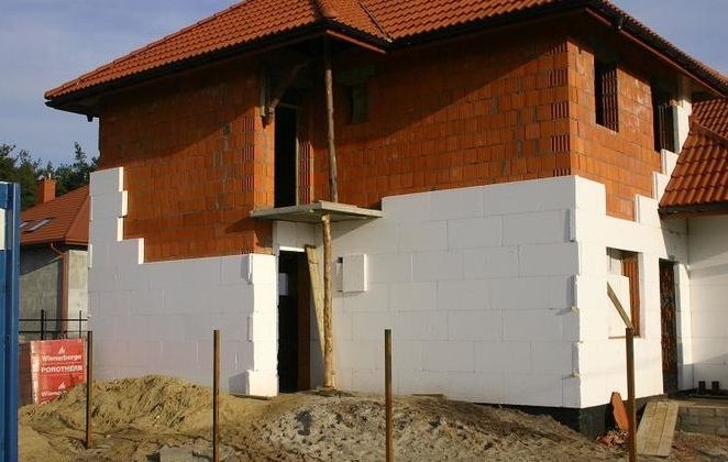 Ocieplanie elewacji domu jednorodzinnego styropianem, widoczne fragmenty muru z cegły. Na pierwszym planie ziemia i tablica reklamowa Wienerberger Porotherm.