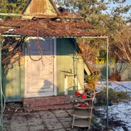 Piotr Dryja - Zrujnowana, drewniana altana z obrośniętymi pnączami, częściowo zasypana śniegiem. Widoczne uszkodzenia dachu i konstrukcji. W tle drzewa i zabudowania.