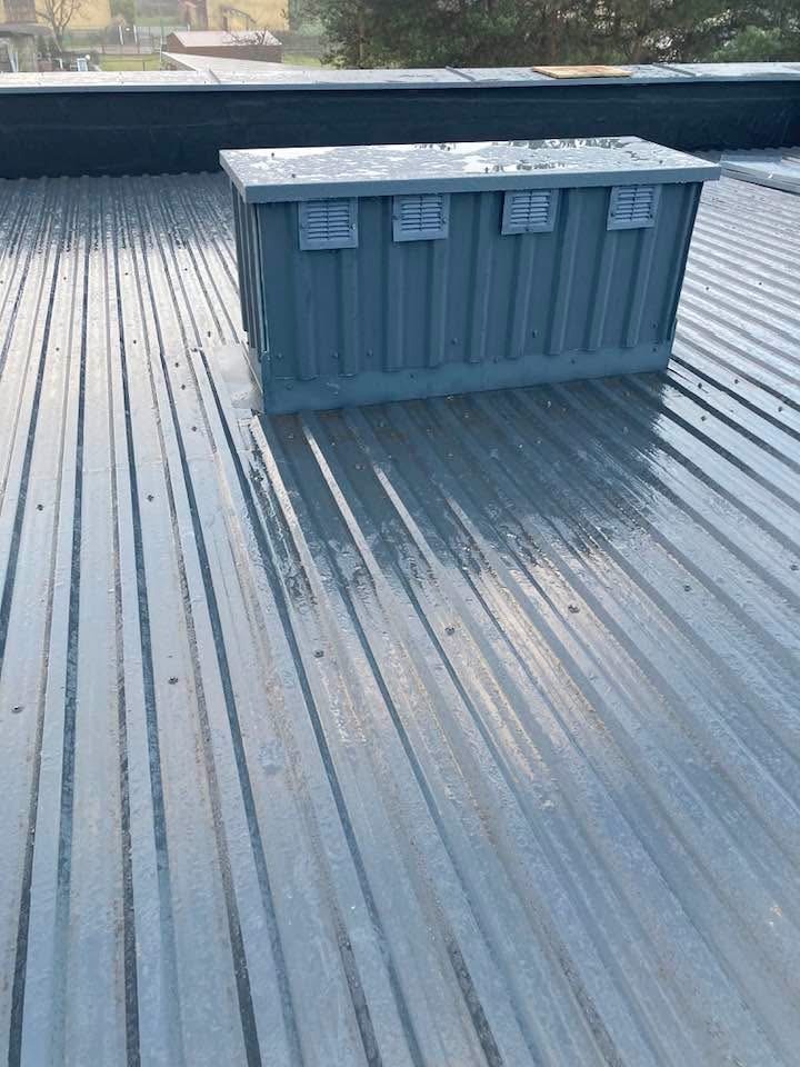 Szara blacha trapezowa na dachu z zamontowaną szarą skrzynką wentylacyjną z czterema otworami wentylacyjnymi. Widoczne mocowania blachy. Dach lekko mokry.