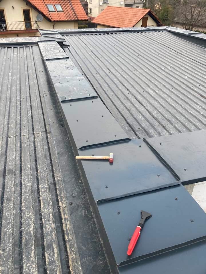 Montaż obróbek blacharskich na dachu z blachy na posesji w Bobrku. Widoczny młotek i nóż dekarski. Obróbki w kolorze grafitowym.