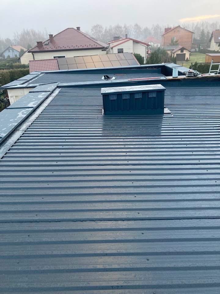 Szary dach z blachy trapezowej z elementami wykończeniowymi i kominem wentylacyjnym na tle zabudowy jednorodzinnej w pochmurny dzień.