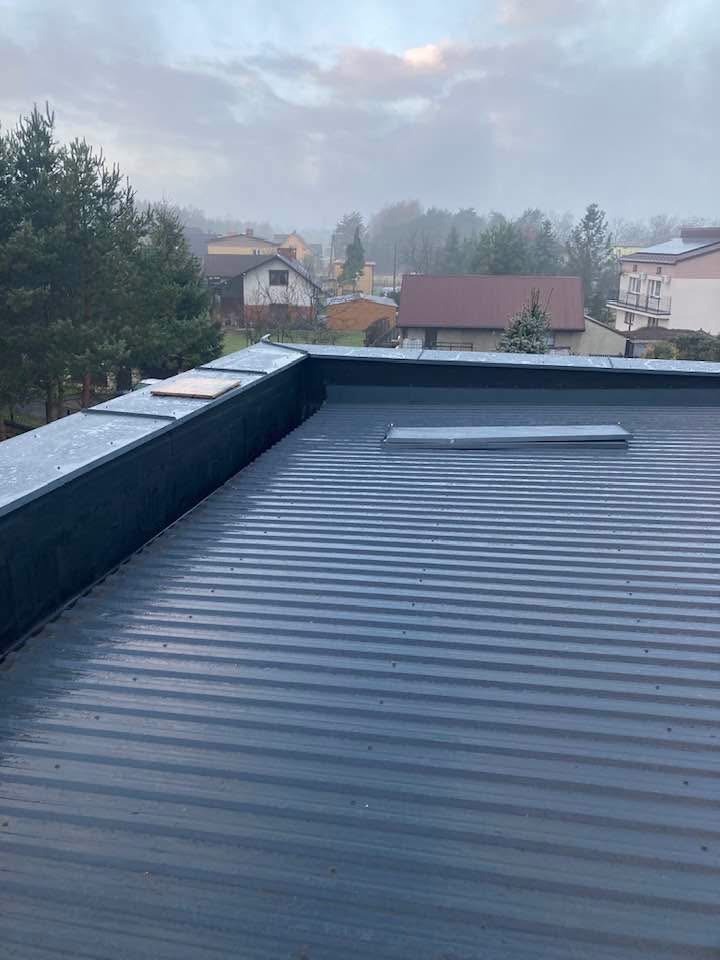 Szary, blaszany dach płaski z obróbkami dekarskimi, widok z góry na tle zabudowy jednorodzinnej w pochmurny dzień. Widoczne detale łączeń blachy.