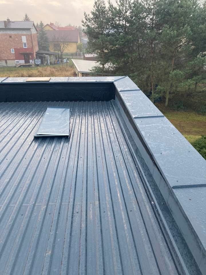 Szary dach z blachy trapezowej z obróbkami blacharskimi, widok z góry na tle zabudowań i drzew. Fragment okna dachowego. Lekko pochmurna pogoda.