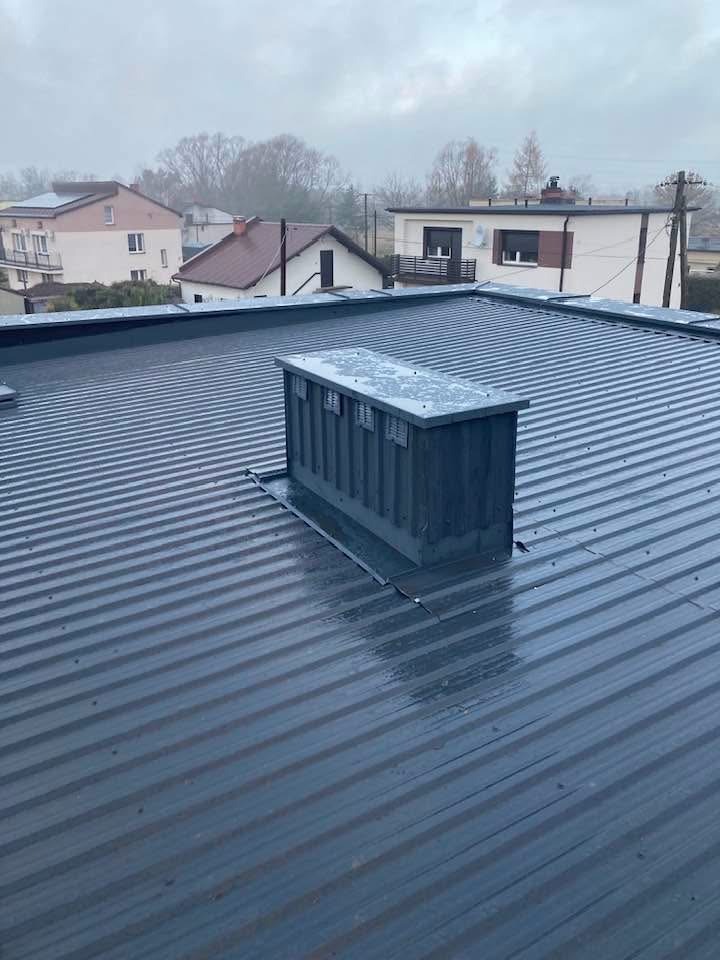 Szary dach z blachy trapezowej z kominem wentylacyjnym na tle zabudowy jednorodzinnej w pochmurny dzień. Widoczne ślady wilgoci na powierzchni dachu.