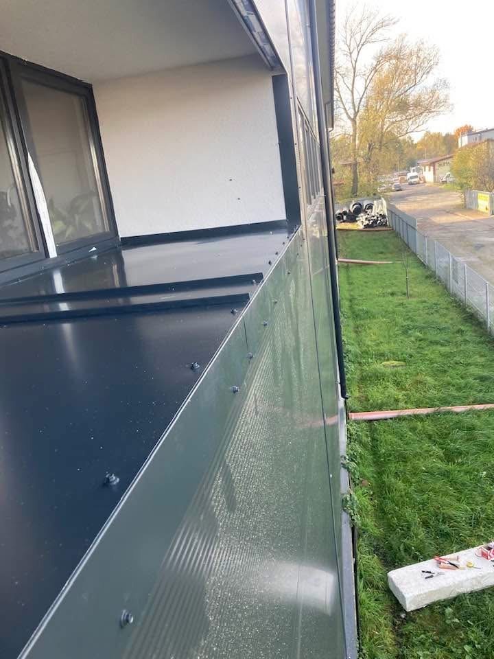 Zamontowana, ciemna blacha na balkonie z widocznymi śrubami mocującymi. W tle trawnik i elementy otoczenia budynku. Ujęcie z boku, podkreślające detale montażu.