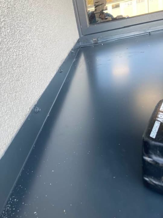 Szczegółowe ujęcie nowej, grafitowej obróbki blacharskiej okna na tle jasnego tynku zewnętrznego budynku. Widoczne ślady po wierceniu i narzędzie pracy.