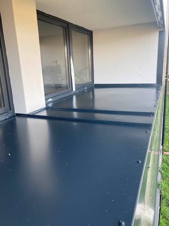 Wykończony balkon z ciemną, metalową podłogą i nowoczesnymi oknami. Widoczne detale łączeń blachy i fragment zieleni w tle. Minimalistyczny design.