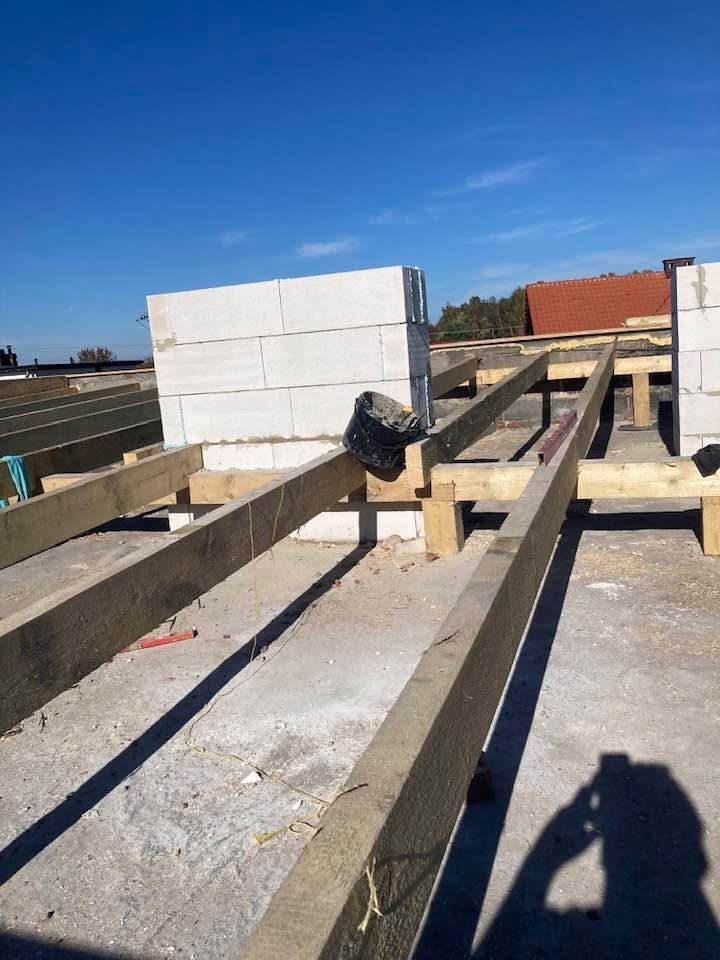 Wznoszenie ścian z betonu komórkowego na drewnianej konstrukcji stropu. Widoczny fragment dachu sąsiedniego budynku i błękitne niebo.