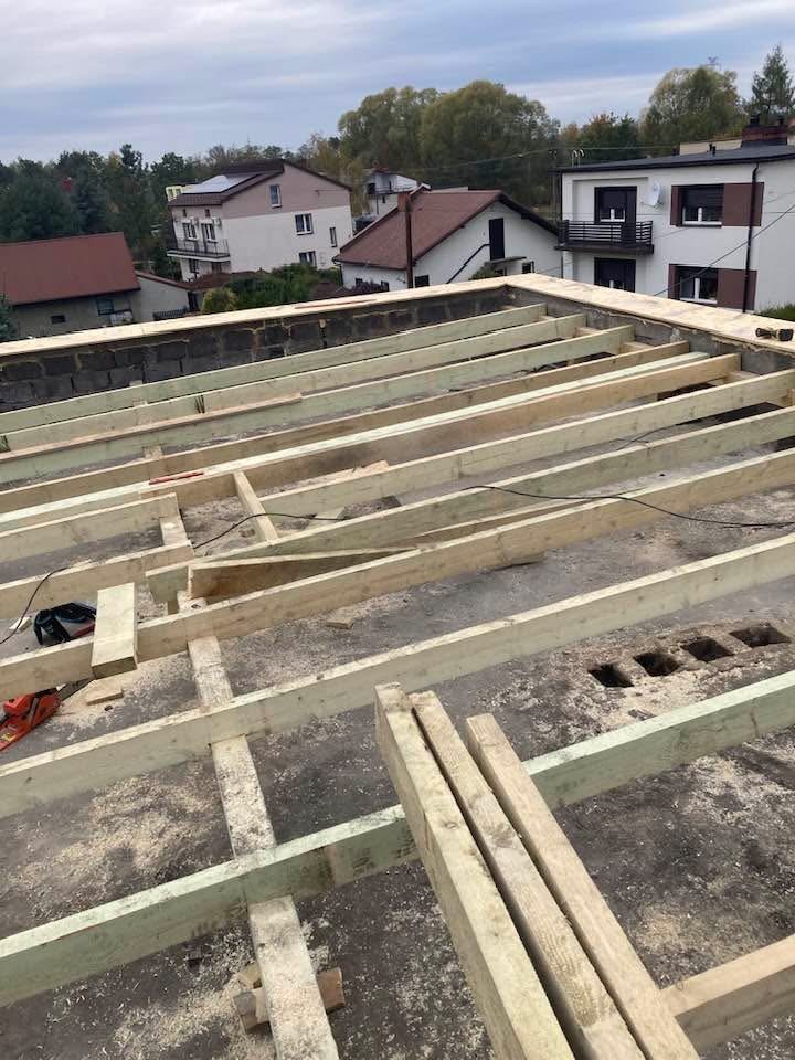 Wiązanie drewnianych belek na betonowym stropie budynku w trakcie budowy. Widoczne narzędzia i otoczenie budowy. Ujęcie z góry, w tle zabudowa jednorodzinna.