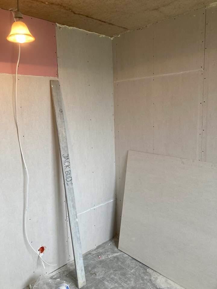 Wnętrze w trakcie wykańczania: ściany obłożone płytami gipsowo-kartonowymi, widoczna poziomica oparta o ścianę, surowy beton na podłodze, lampa wisząca.