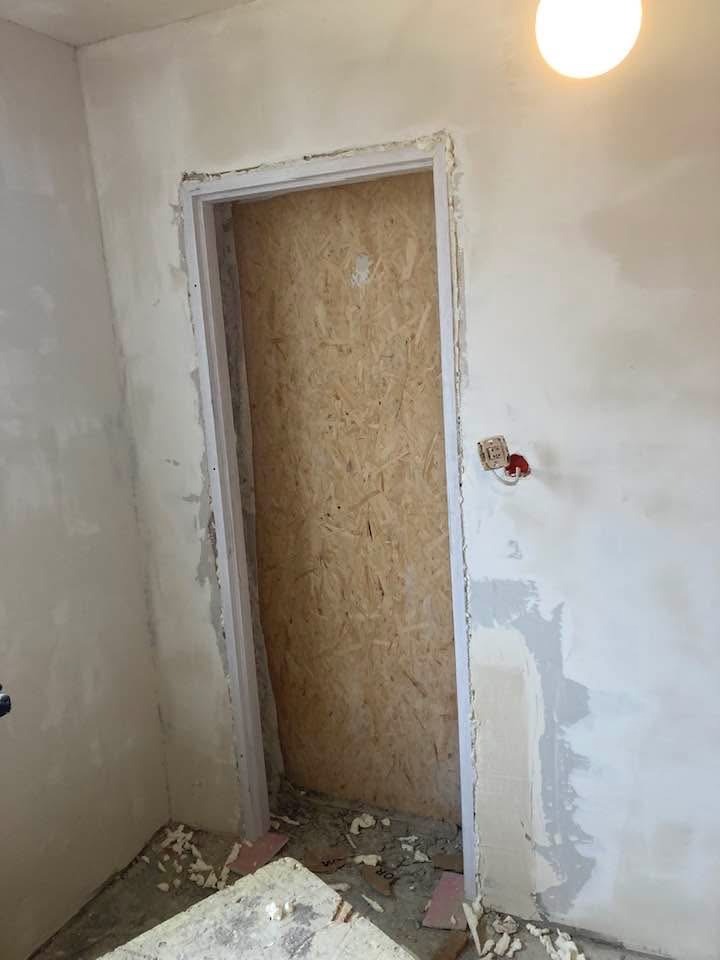 Przygotowany otwór drzwiowy z obramowaniem w surowym stanie, zabezpieczony płytą OSB. Ściany w trakcie wykańczania, widoczne ślady po zaprawie.