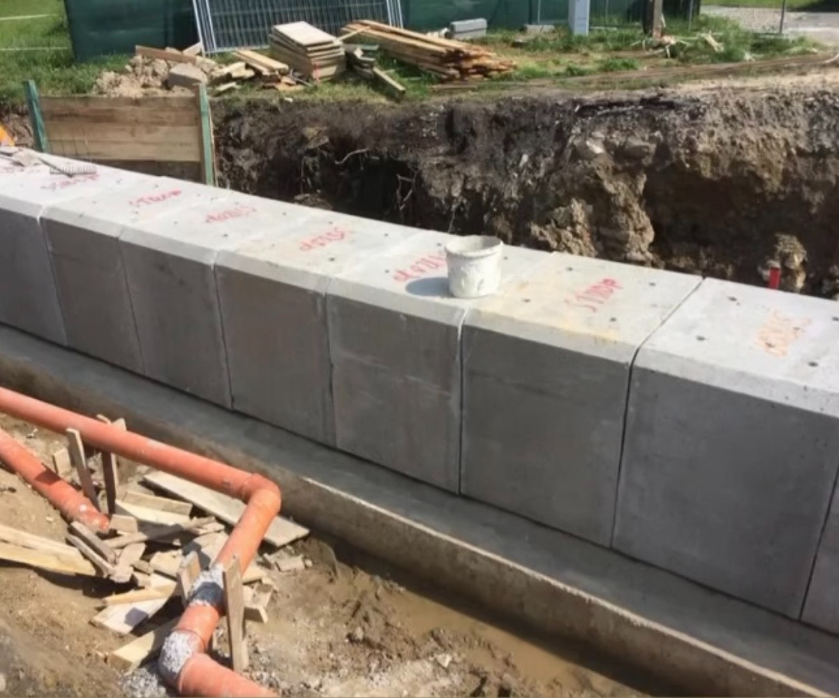 Wyrównane betonowe bloki fundamentowe z widocznymi oznaczeniami, ułożone w wykopie. Pomarańczowe rury i drewniane elementy w pobliżu. Wykopana ziemia w tle.