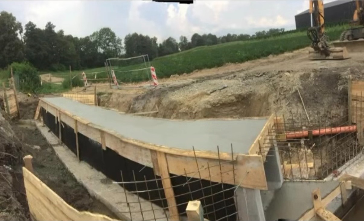 Wylany fundament z szalunkiem i izolacją pionową, widoczna rura kanalizacyjna. W tle krajobraz z zielonym polem i koparka.