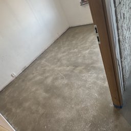 Kacper Starmach - Wnętrze po wylewce samopoziomującej, widoczne ślady po wałku odpowietrzającym. Ściany białe, okno z drewnianą ramą, lampa wisząca. Ujęcie zza futryny.