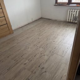 Kacper Starmach - Ukończona podłoga z płytek imitujących drewno w jasnym odcieniu, ułożona w pokoju z oknem i grzejnikiem. Widoczne niedokończone ściany i fragment ościeżnicy.