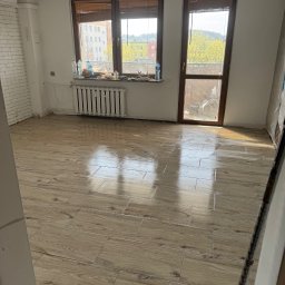 Kacper Starmach - Ułożona podłoga z płytek imitujących drewno w jasnym pokoju z oknem i grzejnikiem. Widoczne niedokończone ściany i sufit.