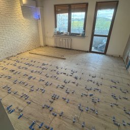 KS-Bud Usługi glazurnicze, remont łazienki, wykończenia - Układanie płytek podłogowych w jasnym pomieszczeniu z ceglaną ścianą. Widoczne niebieskie klipsy poziomujące. Dwa okna z widokiem na zewnątrz.