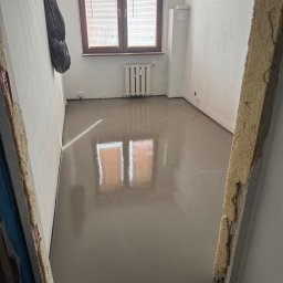 Kacper Starmach - Wylana, gładka posadzka samopoziomująca w jasnym pokoju z oknem i grzejnikiem. Widoczne ślady po szalunku. Na ścianie wisi kurtka.