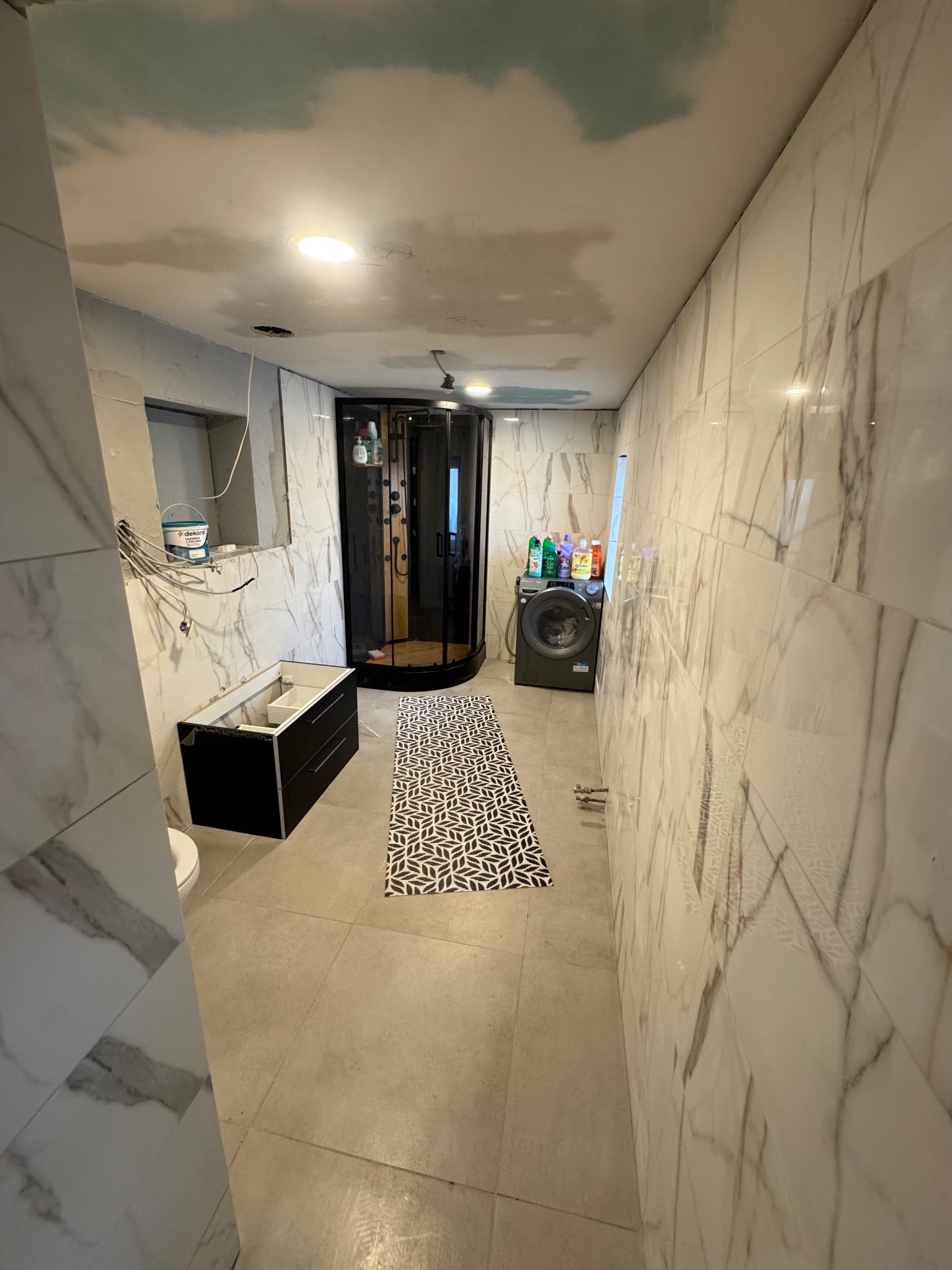 Wnętrze łazienki w trakcie wykańczania: marmurowe płytki, kabina prysznicowa, pralka, niedokończona szafka pod umywalkę, wzorzysty dywanik i sufit z motywem nieba.