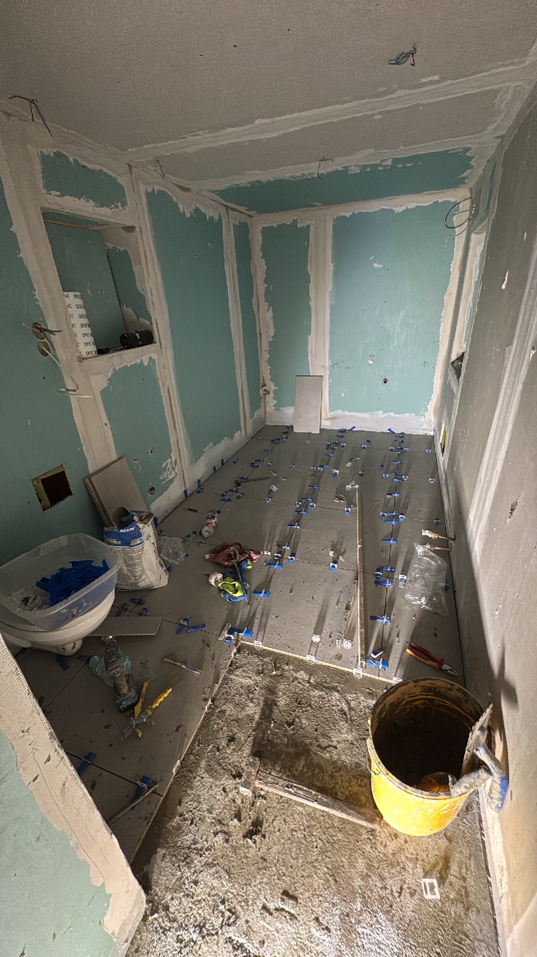 Wnętrze łazienki w trakcie prac wykończeniowych: ułożone płytki podłogowe z krzyżykami dystansowymi, ściany przygotowane pod malowanie, widoczne narzędzia i materiały budowlane.