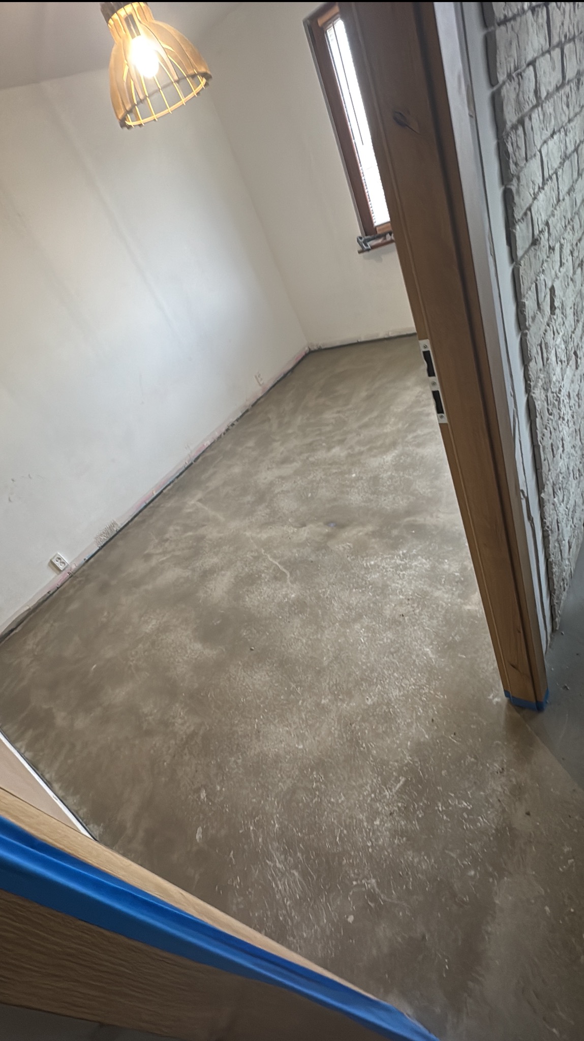 Wnętrze po wylewce samopoziomującej, widoczne ślady po wałku odpowietrzającym. Ściany białe, okno z drewnianą ramą, lampa wisząca. Ujęcie zza futryny.