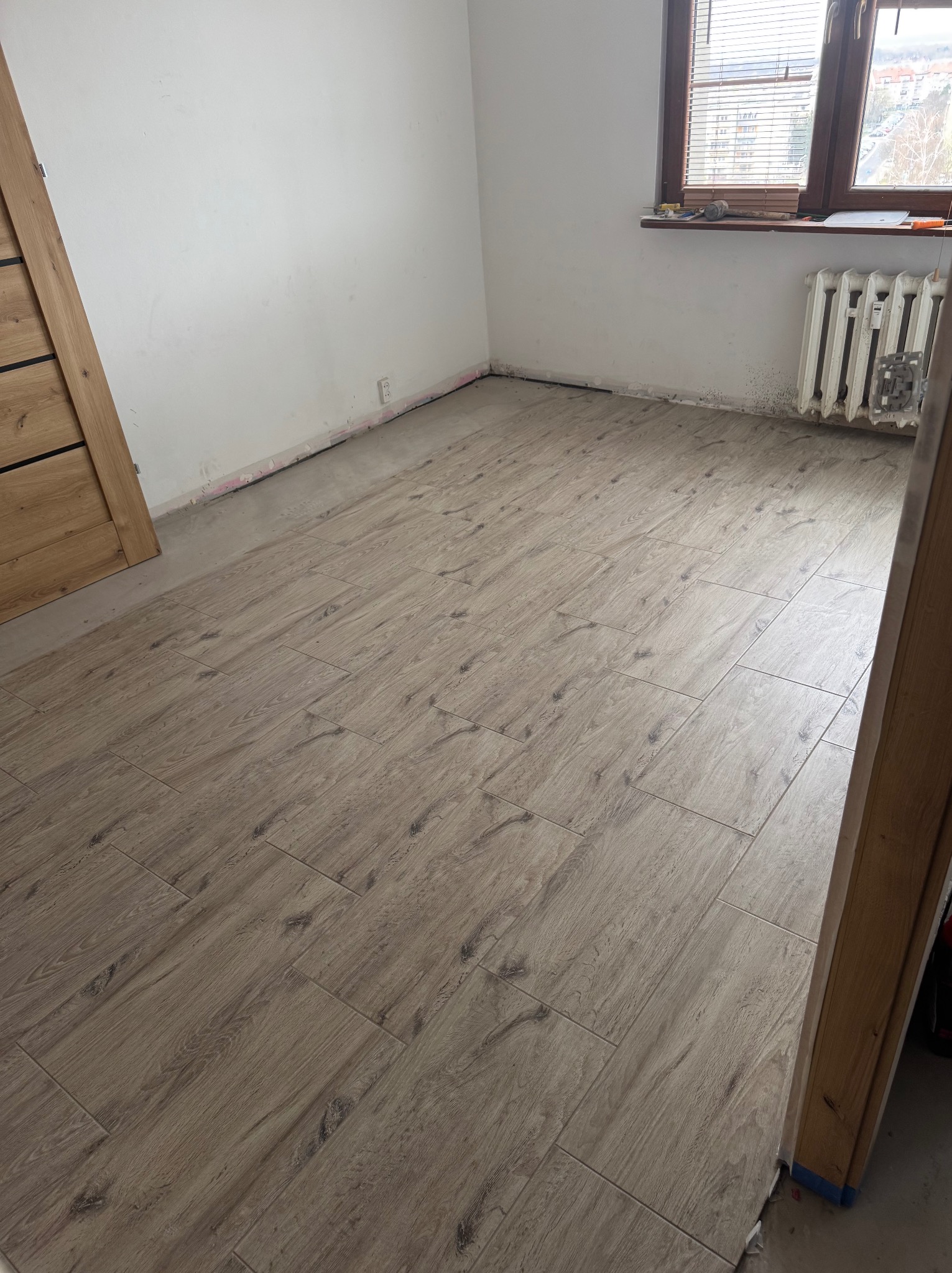 Ukończona podłoga z płytek imitujących drewno w jasnym odcieniu, ułożona w pokoju z oknem i grzejnikiem. Widoczne niedokończone ściany i fragment ościeżnicy.