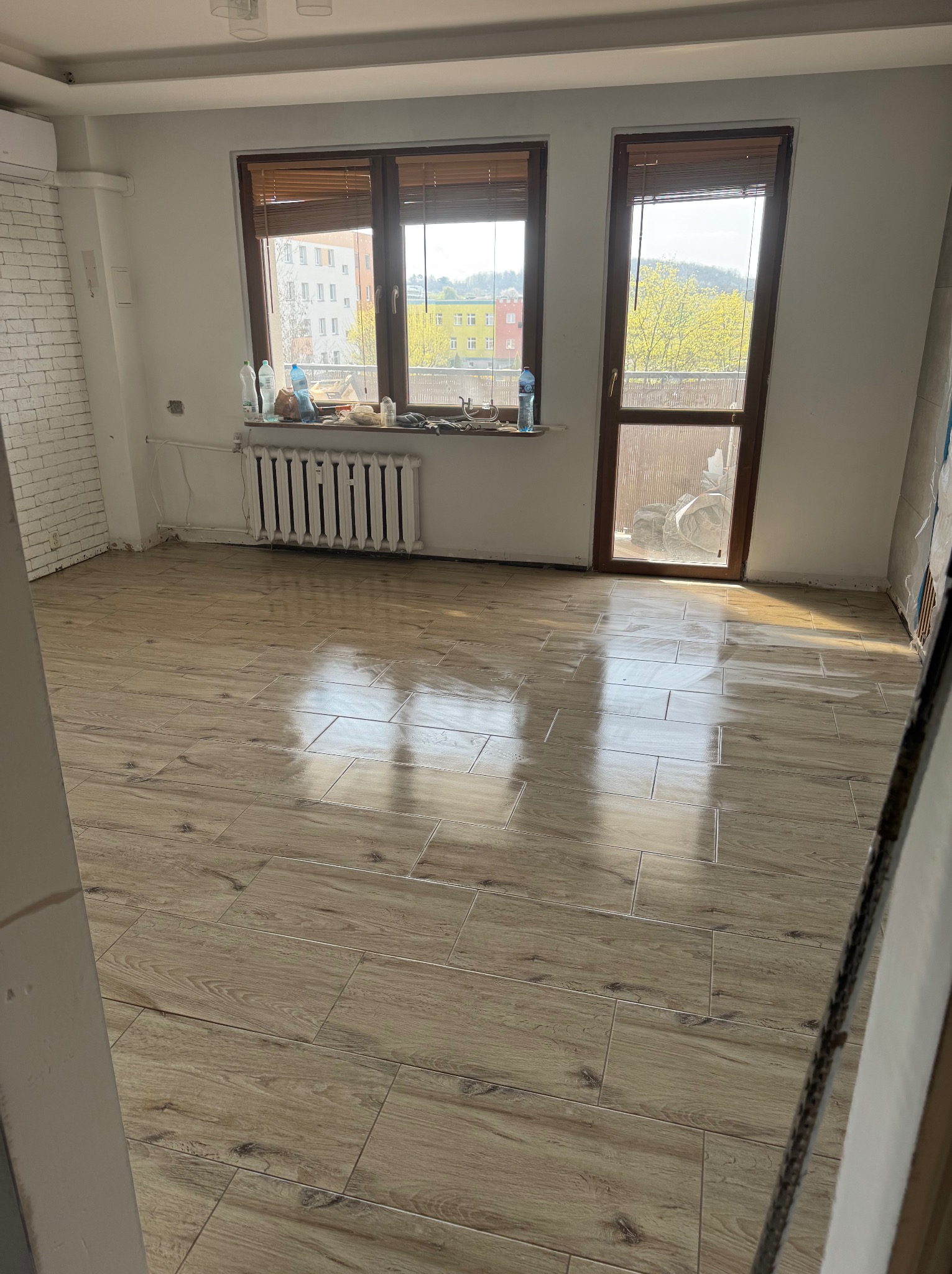 Ułożona podłoga z płytek imitujących drewno w jasnym pokoju z oknem i grzejnikiem. Widoczne niedokończone ściany i sufit.