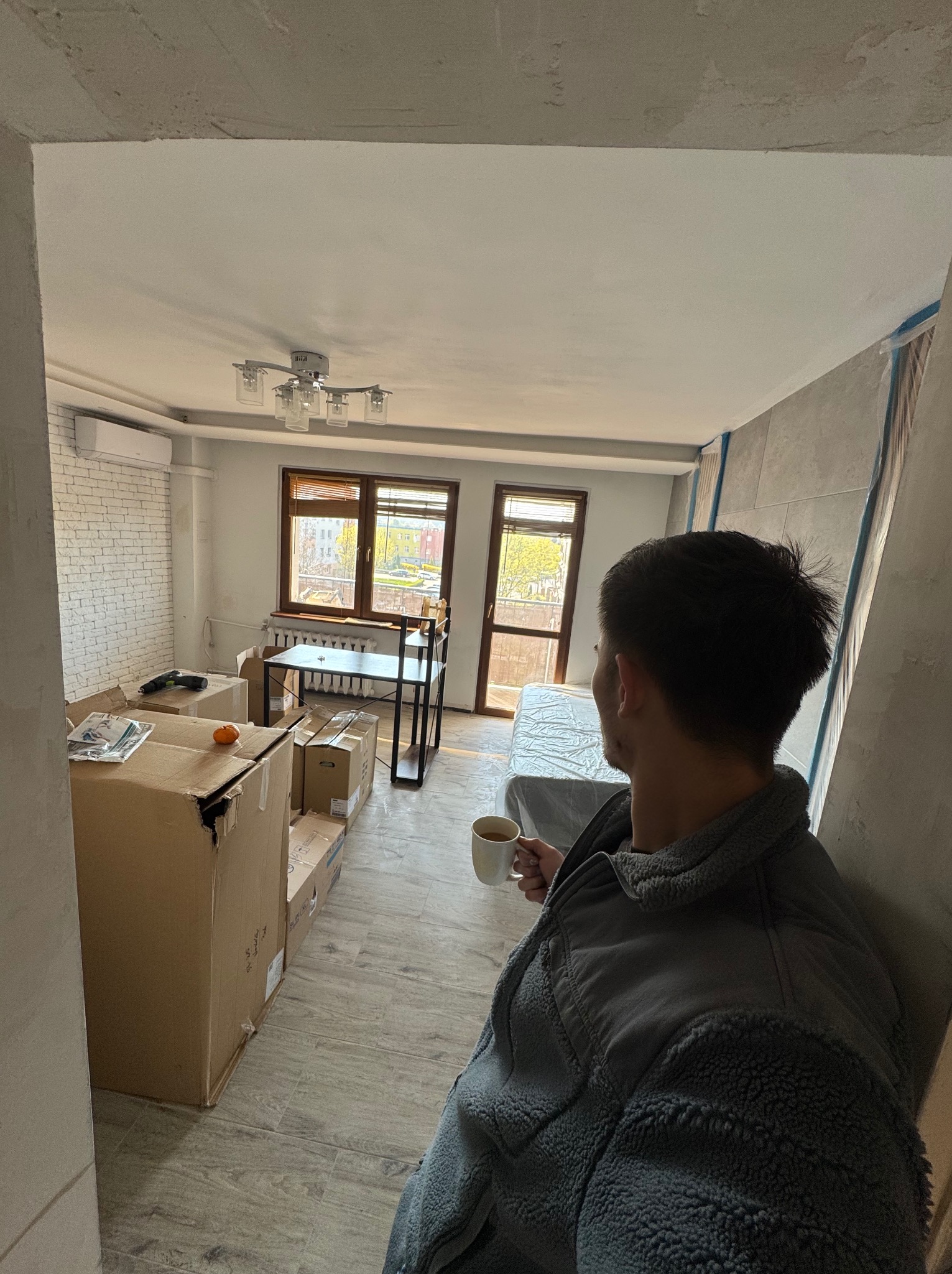 Mężczyzna z kubkiem w dłoni obserwuje wnętrze mieszkania w trakcie prac wykończeniowych. Widoczne kartony, odsłonięta cegła i zabezpieczone ściany.