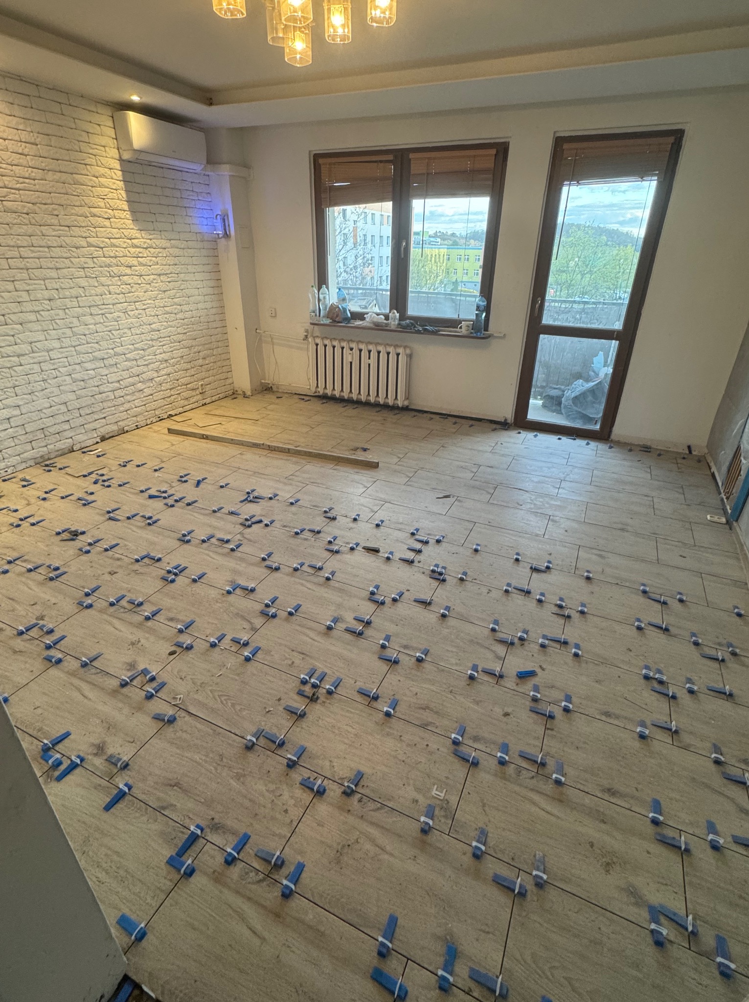 Układanie płytek podłogowych w jasnym pomieszczeniu z ceglaną ścianą. Widoczne niebieskie klipsy poziomujące. Dwa okna z widokiem na zewnątrz.