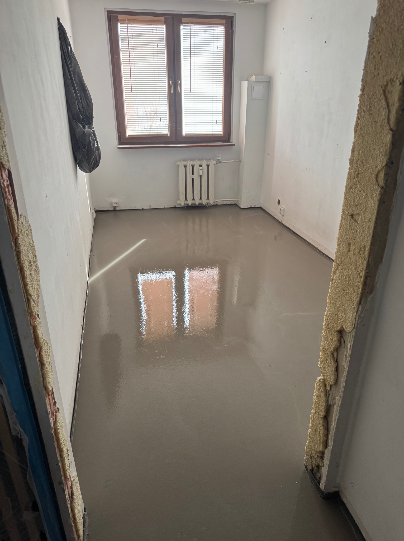 Wylana, gładka posadzka samopoziomująca w jasnym pokoju z oknem i grzejnikiem. Widoczne ślady po szalunku. Na ścianie wisi kurtka.