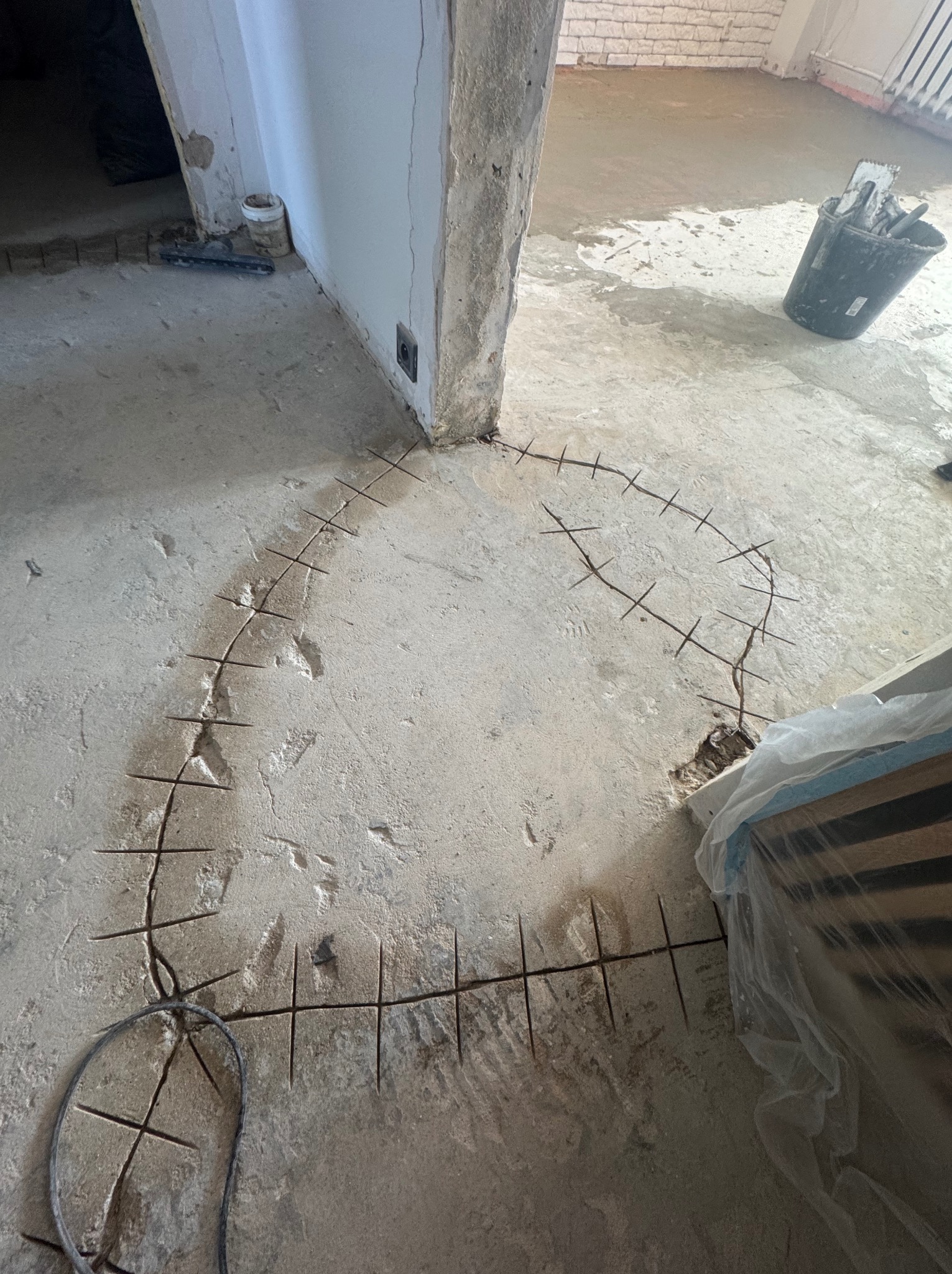 Przygotowanie podłoża pod wylewkę: nacięcia w kształcie łuku na betonowej podłodze, widoczne narzędzia i wiadro. Etap prac wykończeniowych w pomieszczeniu.