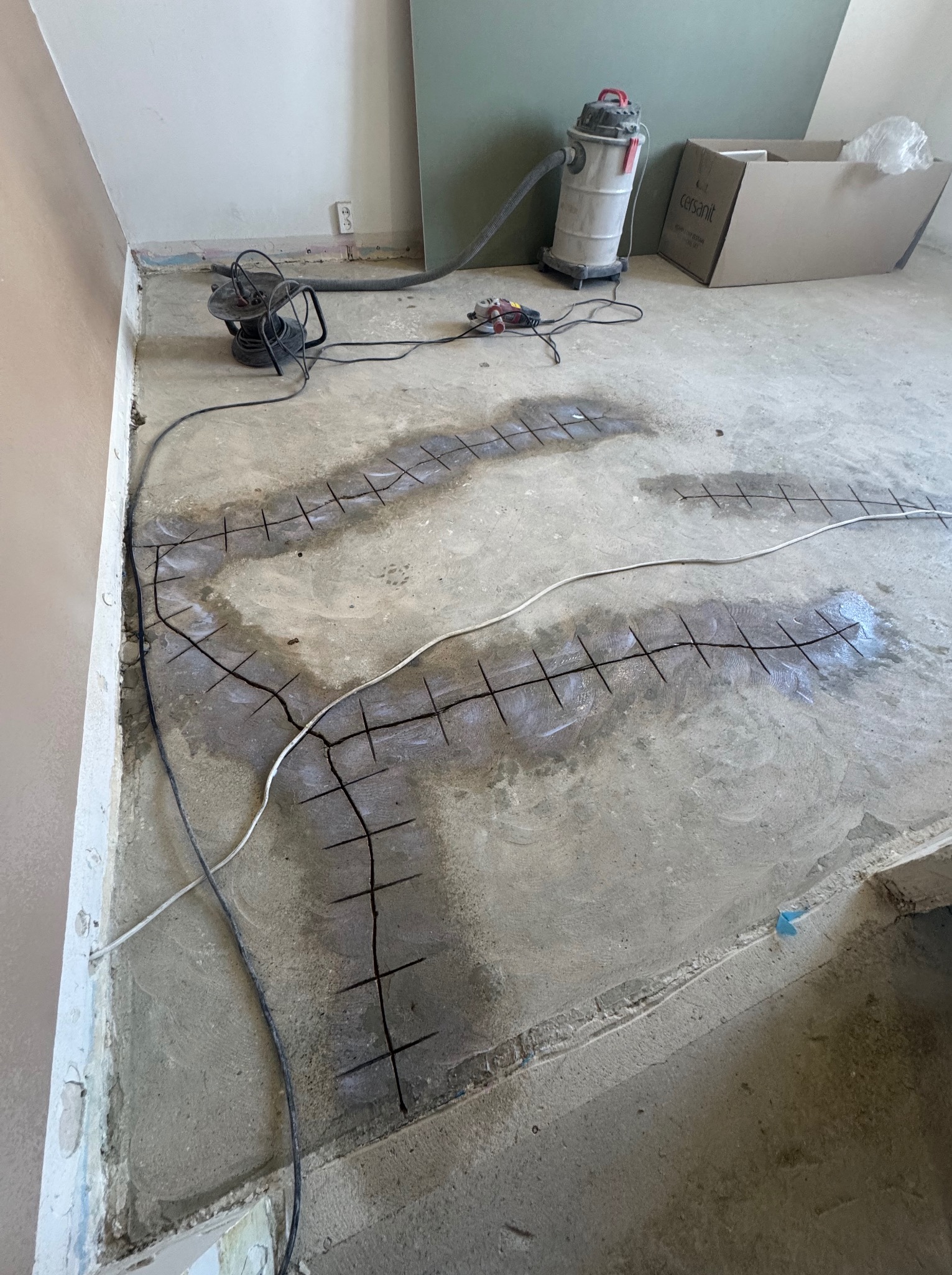 Przygotowanie podłoża pod ogrzewanie podłogowe w Koźlicach. Wycięte rowki w betonie, widoczny odkurzacz przemysłowy i przewody elektryczne. Etap prac przed wylaniem wylewki.