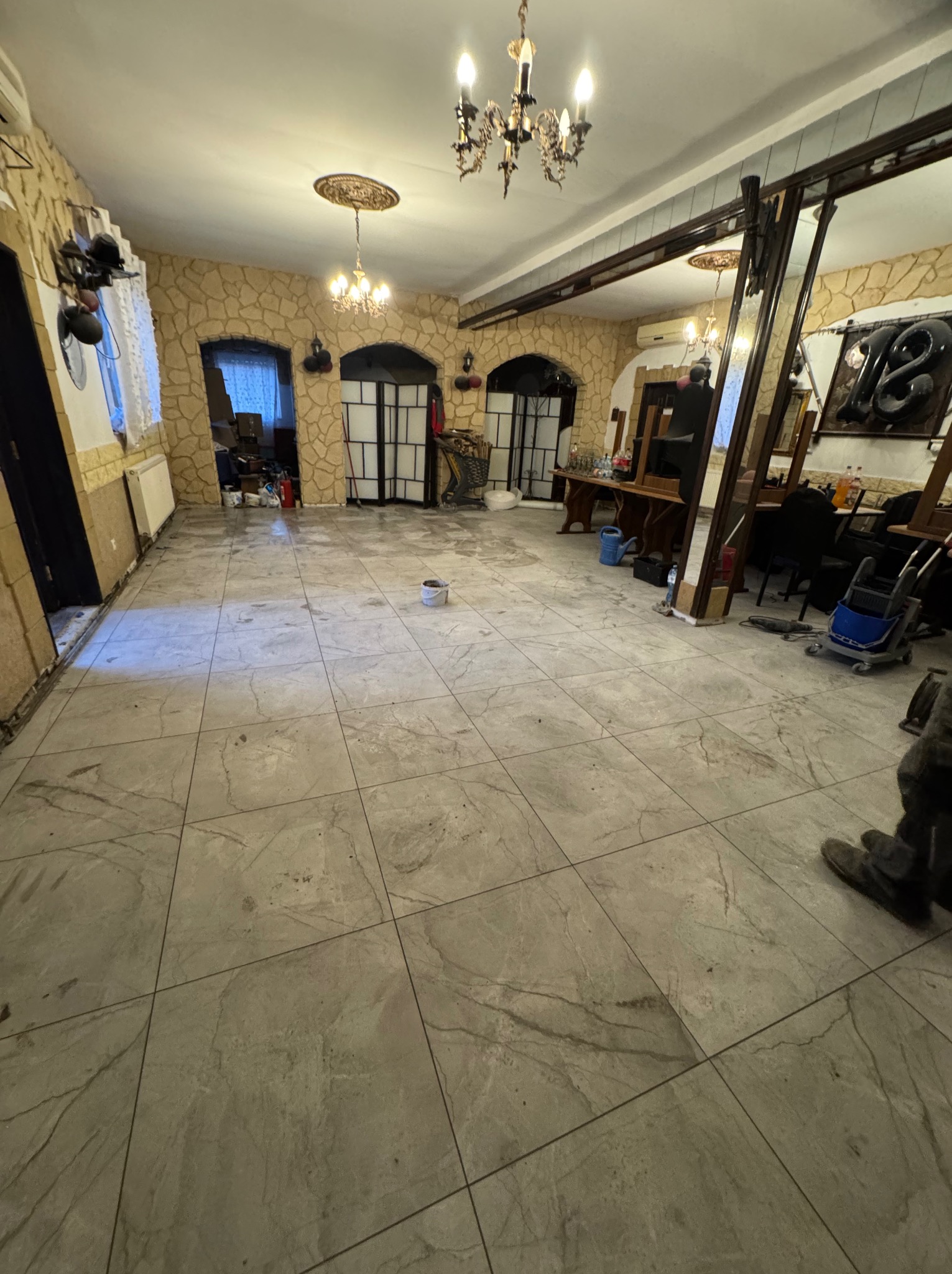 Ułożona, jasnoszara podłoga z płytek imitujących kamień w przestronnym wnętrzu z dekoracyjną ścianą i oświetleniem. Widoczny fragment buta w prawym dolnym rogu.