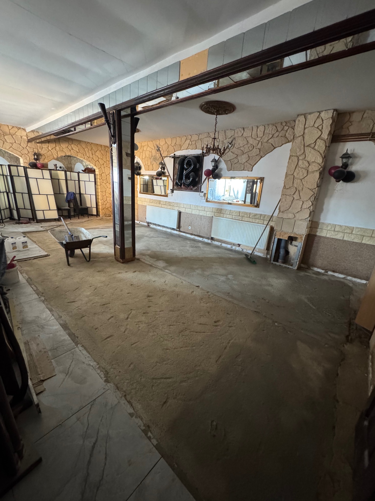 Wnętrze w trakcie prac przygotowawczych do wykończenia podłogi. Widoczna taczka z narzędziami, ściany z dekoracyjnym kamieniem i przygotowane podłoże.