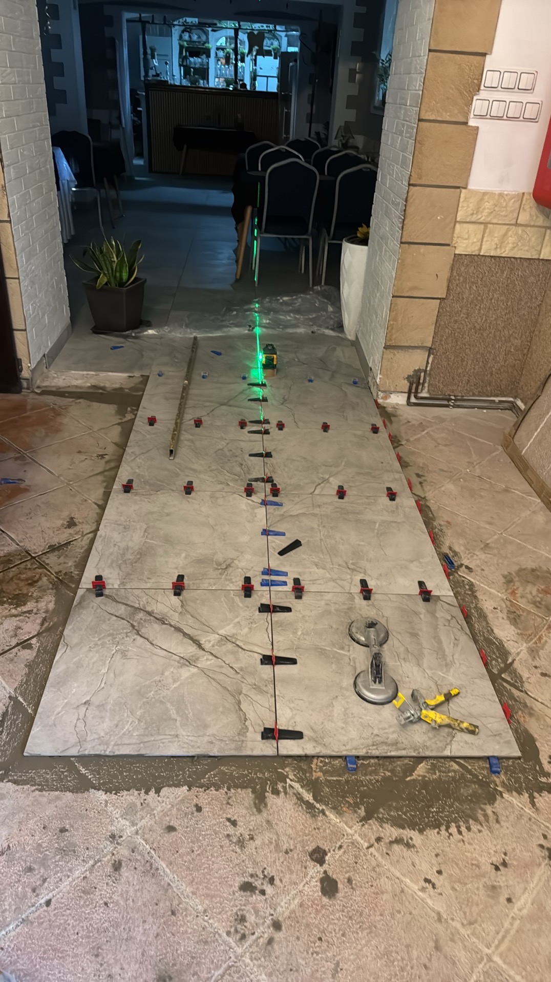 Układanie płytek podłogowych z użyciem poziomicy laserowej i systemu poziomowania. Widoczne narzędzia glazurnicze i świeżo nałożona zaprawa.