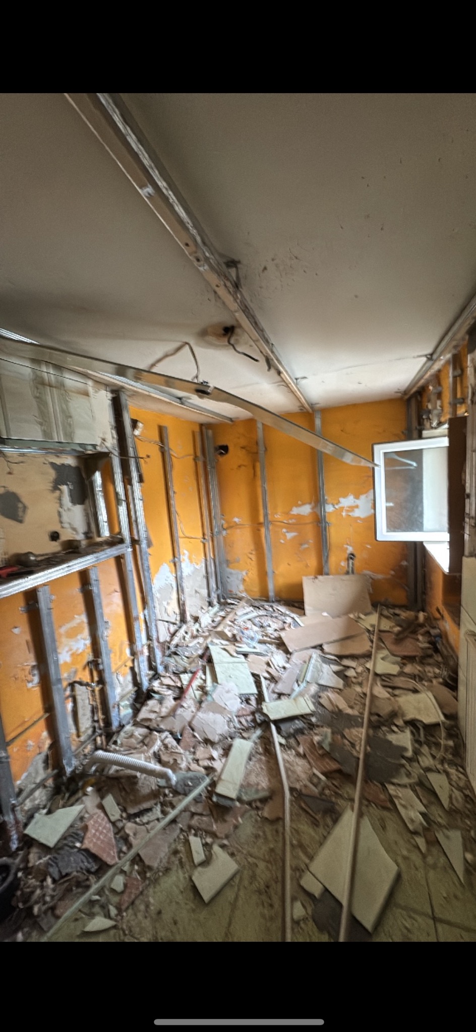 Wnętrze w trakcie demontażu: pomarańczowe ściany z odsłoniętymi instalacjami, metalowe profile, gruz na podłodze. Widoczne okno i fragment sufitu z elementami konstrukcyjnymi.