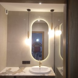 Usługi remontowe Łukasz Majewski - Nowoczesna łazienka z owalnym lustrem podświetlanym LED, wiszącą umywalką na blacie z marmurowym wzorem i złotą armaturą. Widoczne lampy wiszące i odbicie osoby w lustrze.