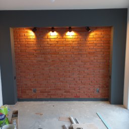 Usługi remontowe Łukasz Majewski - Ceglana ściana z oświetleniem punktowym w ramie pomalowanej na ciemny kolor. Na podłodze widoczne narzędzia i materiały budowlane.