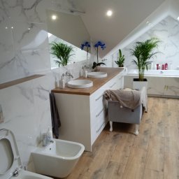 Usługi remontowe Łukasz Majewski - Jasna łazienka na poddaszu z drewnianą podłogą, marmurowymi płytkami i białymi meblami. Umywalki nablatowe, bidet i toaleta w nowoczesnym stylu. Rośliny doniczkowe dodają świeżości.