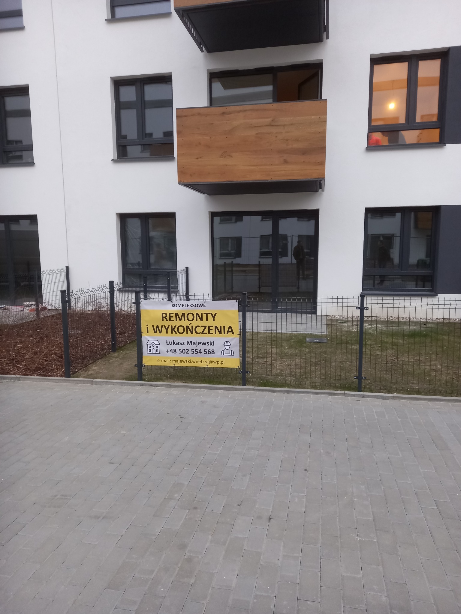 Nowoczesny budynek z balkonami i oknami w szarych ramach. Na pierwszym planie żółta tablica informacyjna z ofertą remontów i wykończeń.