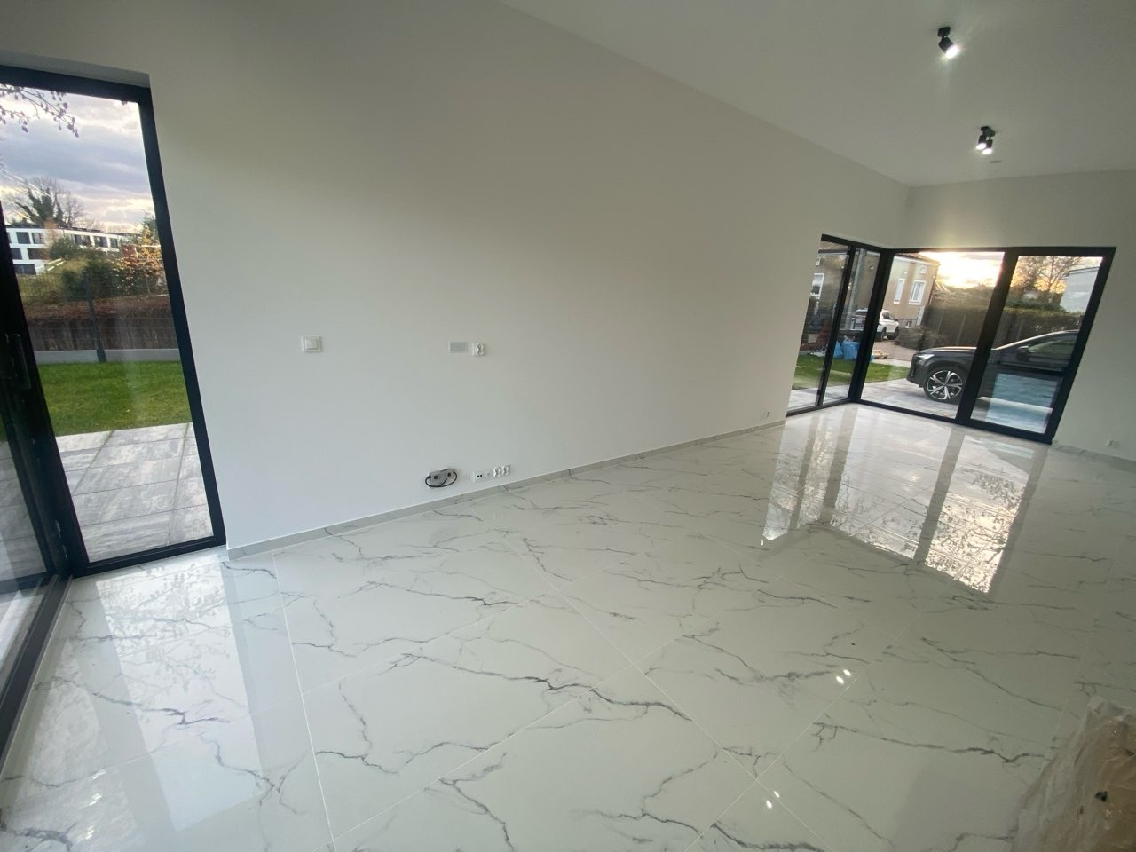 Jasne, przestronne wnętrze po remoncie: białe ściany, marmurowe płytki podłogowe z szarymi żyłami, duże okna z czarnymi ramami wpuszczające światło dzienne.