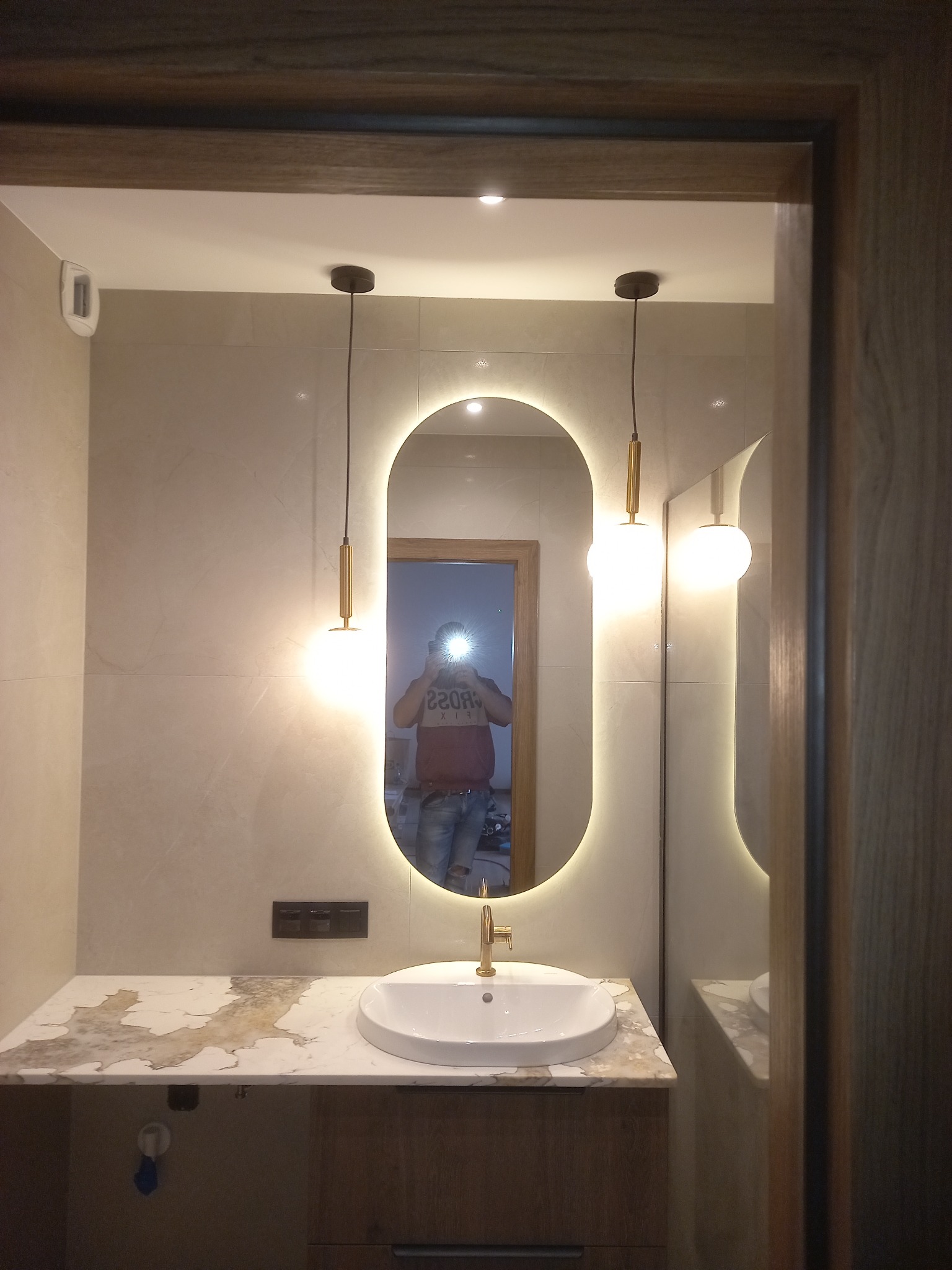 Nowoczesna łazienka z owalnym lustrem podświetlanym LED, wiszącą umywalką na blacie z marmurowym wzorem i złotą armaturą. Widoczne lampy wiszące i odbicie osoby w lustrze.