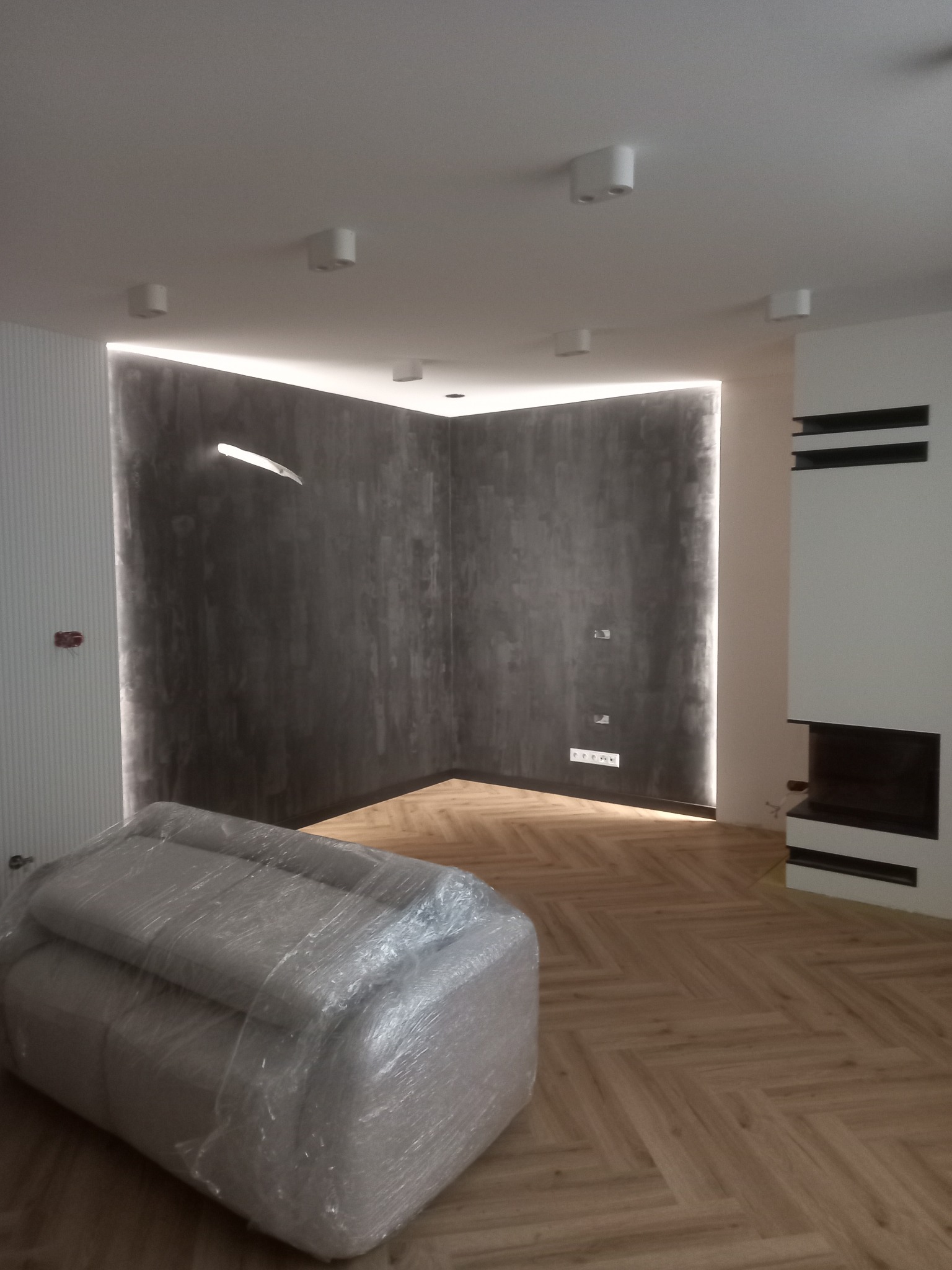 Nowoczesne wnętrze po remoncie: parkiet w jodełkę, ściana z oświetleniem LED, biała zabudowa kominkowa. Sofa zabezpieczona folią ochronną.